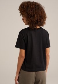 Černé bavlněné tričko s krátkými rukávy, uvolněný střih a žebrovaný výstřih. Viditelné švy na zadní straně, spojené s olivově zelenými kalhotami.