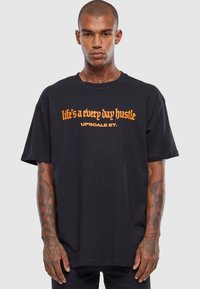 Black cotton t-shirt with loose fit, featuring bold orange text "life's a every day hustle" and "UPSCALE ST." in decorative font.