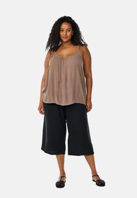 Braunes, ärmelloses Top mit gerafftem Ausschnitt, kombiniert mit schwarzen, weit geschnittenen Cropped-Hosen. Das Model trägt schwarze, schnürbare Sandalen. Glatte Stoffstruktur.