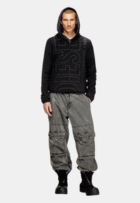 Haut à capuche noir zippé avec un logo imprimé bien en vue, associé à un pantalon cargo gris surdimensionné et des bottes noires. Texture lisse, coupe décontractée.