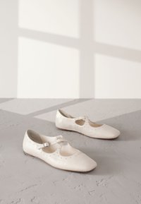 Fehér bőr Mary Jane cipők kerek orral, díszítő szegecselt részletekkel az oldalukon, és egy állítható pánttal.