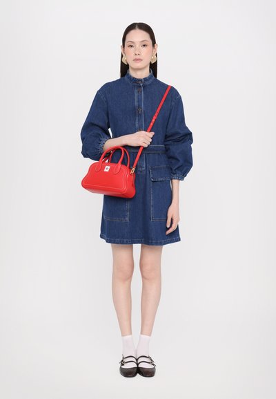 Robe en denim avec des manches bouffantes, taille cintrée, poches avant. Le modèle tient un petit sac à main en cuir rouge avec des poignées doubles et une bandoulière.