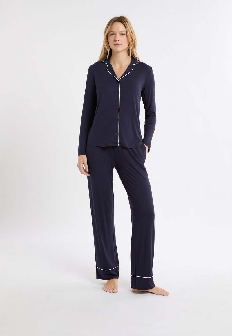 Set di pigiama blu navy con maniche lunghe e pantaloni. Presenta piping bianco a contrasto lungo il colletto, i bottoni e le estremità dei pantaloni. Tessuto morbido e liscio.