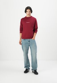 Burgundy long-sleeve sweatshirt with white embroidered text, paired with light blue baggy jeans and black sneakers. Plain background.