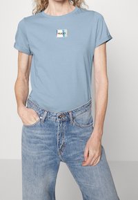 Žena v světle modrém tričku s vy rolled-sleeve a holografickou nášivkou "HUGO" a v světle modrých džínech s vysokým pasem, s jednou rukou v kapse.
