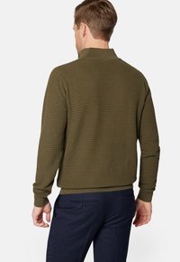 Olive green pulower z dzianiny waffle z stójką, dopasowanymi mankietami i prostym dołem, w połączeniu z ciemnymi spodniami. Pokazano widok z tyłu.