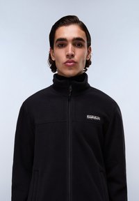 Black fleece jacket with a high collar, front zip, and side pockets. Features a logo patch on the chest and textured fabric.