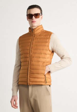Gilet matelassé ambre avec un col haut, dotée de poches zippées et d'une texture lisse, superposé sur un pull beige clair.