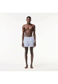 Boxer shorts con base bianca, caratterizzati da un motivo a foglie verdi, cintura elastica e vestibilità ampia, indossati da un uomo in piedi.