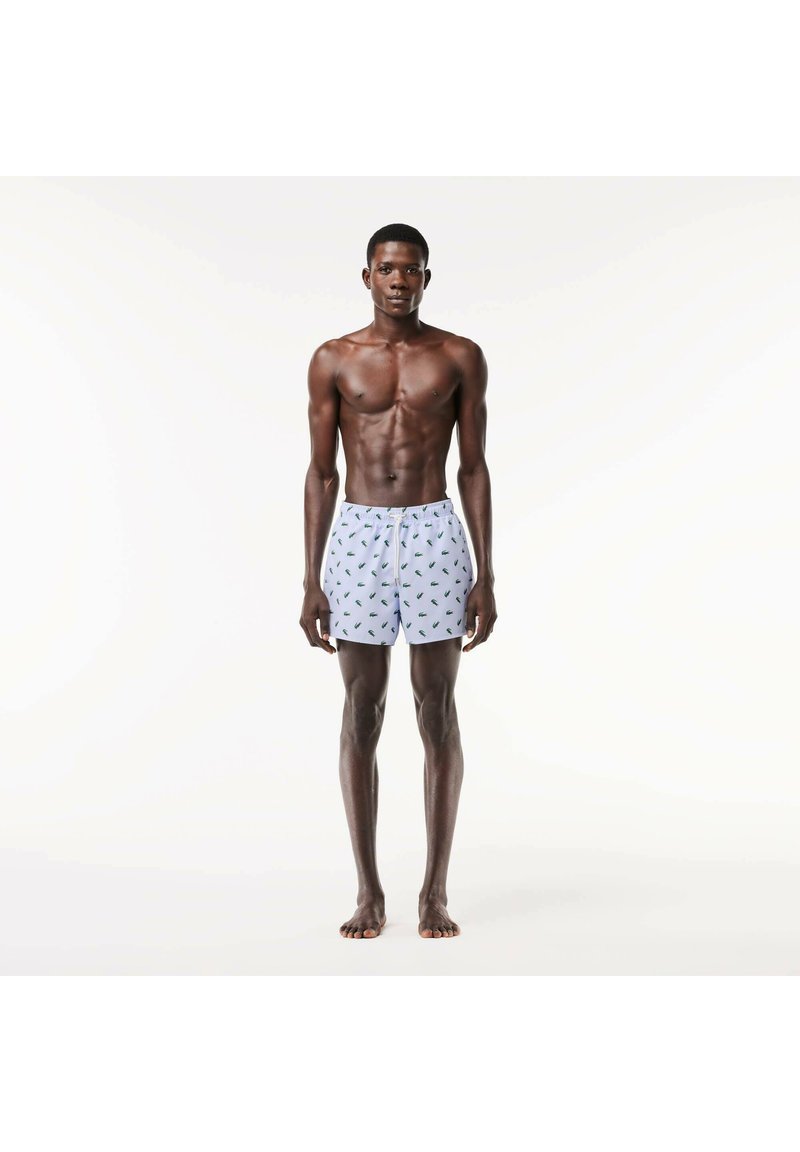 Boxer shorts con base bianca, caratterizzati da un motivo a foglie verdi, cintura elastica e vestibilità ampia, indossati da un uomo in piedi.