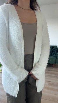 Hvit fluffy cardigan med åpen front, lange ermer og ribbede mansjetter, sammen med en beige topp og mønstrede grå bukser.