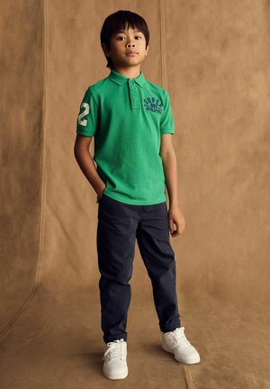 Grön polo shirt med "Super Dry Athletics" tryckt, nummer 2 på ärmen, i kombination med mörka byxor och vita sneakers mot en neutral bakgrund.