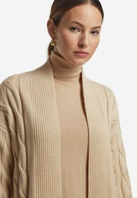 Beige stickad cardigan med ribbat öppet fram och texturerade kabelmönster. Underneath en matchande beige polotröja.