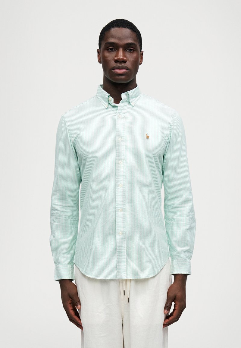 Man i ljusgrön och vit randig skjorta med knappar och vita byxor med dragsko som står mot en enkel vit bakgrund.