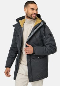 Chaqueta aislante gris oscuro con capucha forrada de piel sintética, bolsillos con cremallera y solapa abotonada. Cremallera frontal y cordón en la capucha.