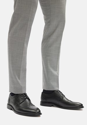 Czarne skórzane buty na dress code z detalami brogue, o gładkim wykończeniu, wiązanym fasonie i zaokrąglonym nosku, zestawione z szarymi spodniami w kratę.