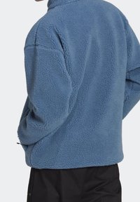 Ljust blå fleecepolo med hög krage, strukturerad yta och avslappnad passform. Har sidofickor och elastisk fåll i nederkant.