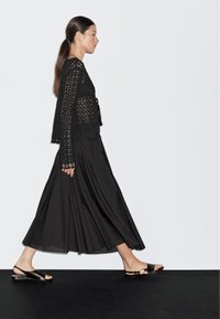 Top nero all'uncinetto con maniche lunghe, abbinato a una gonna nera fluida e sandali bassi. L'outfit presenta contrasti di texture e una vestibilità ampia.