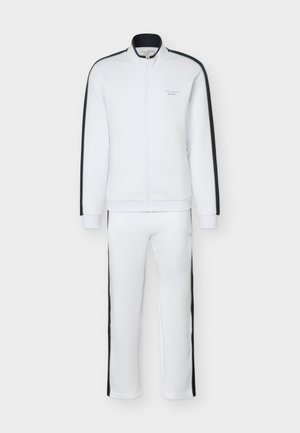 Survêtement blanc zippé avec des rayures noires sur les manches et les jambes, avec le logo "Ted Baker Sport" sur la poitrine et la cuisse.