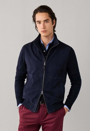 Joven con chaqueta azul marino con cremallera sobre una camisa azul y pantalones burdeos, de pie con las manos en los bolsillos contra un fondo liso.