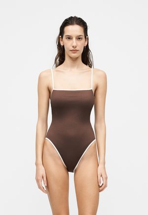 Femme aux longs cheveux foncés portant un maillot de bain une pièce marron avec des bordures blanches, debout devant un fond clair uni.