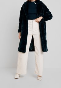 Abrigo de piel sintética verde oscuro con gran cuello, llevado sobre una blusa de punto oscura, combinado con pantalones de pana crema de pierna ancha y tacones beige.
