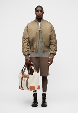 Homme debout portant une veste bomber beige, un pull gris, un short marron, des bottes foncées, tenant un grand sac fourre-tout blanc et marron avec un logo.