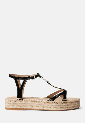 Sandale espadrille en cuir verni noir avec bride à la cheville, plateforme en jute tressé et détails entrecroisés. Semelle plate avec bord de corde texturée.