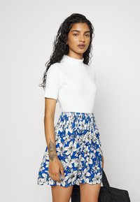 Top branco de manga curta combinado com uma saia azul floral com cintura franzida e intrincados padrões de flores brancas. Bolsa preta segurada numa mão.
