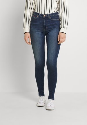 Kobieta w ciemnoniebieskich obcisłych dżinsach, białych trampkach i białym swetrze z długim rękawem z czarnymi pionowymi paskami.