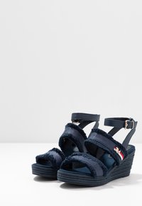 Sandali a zeppa blu navy con cinturini in tessuto sfilacciato, cinturino alla caviglia con fibbia, suola texturizzata e un accento in rosso e bianco contrastante sul lato.