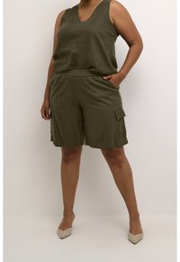 Kvinde iført en olivengrøn ærmeløs top og matchende knælange cargoshorts med beige spidse højhælede sko, stående med den ene hånd i lommen.
