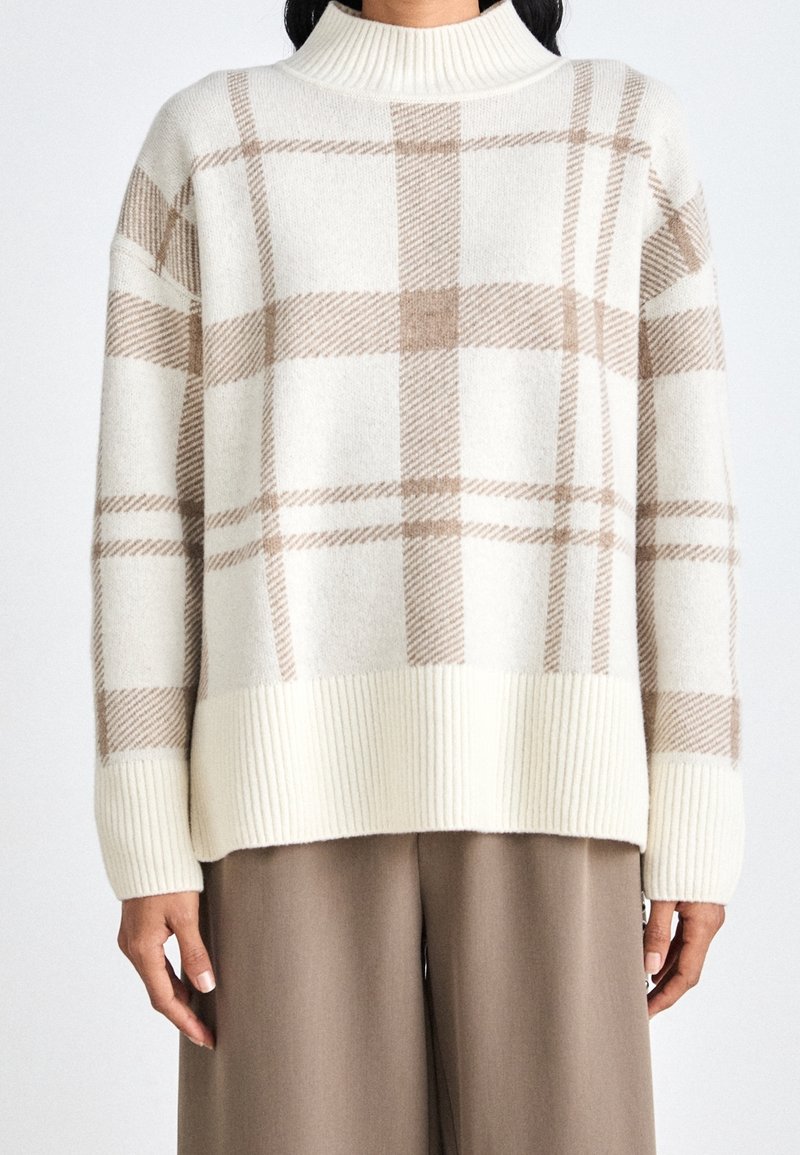 Pull crème avec un motif à carreaux beige, col et poignets côtelés. Texture douce avec une coupe décontractée, associée à un pantalon marron.