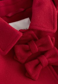 Roter Filzmantelkragen mit zwei auffälligen Schleifen. Der Stoff ist weich mit einer glatten Textur und sichtbaren Nähten an den Kanten.