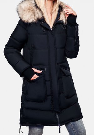 Femme portant une parka matelassée noire avec capuche bordée de fourrure, poches latérales et passe-pouces, avec un jean clair, debout devant un fond blanc.