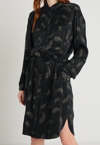 Svart knäppskjortklänning med insnoffsad midja, som har ett fjädermönster och sidslitsar. Tillverkad av ett mjukt, lätt material.