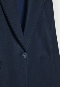 Marineblå uldblazer med en notch-krave, enkeltknapslukning og to forlommer. Blød struktur med subtile stofdetaljer.