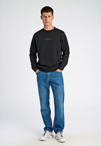 Musta collegepaita, jossa on "LINDBERGH"-teksti, yhdistettynä sinisiin farkkuihin ja valkoisiin tennareihin. Asu korostaa rentoa kaupunkityyliä.