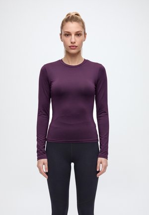 Une femme porte un haut de sport à manches longues couleur prune et un legging noir, sur un fond uni clair.