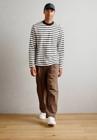 Chemise à rayures à manches longues blanche et bleu marine, pantalon marron et baskets blanches. Le modèle porte une casquette grise et se tient sur un sol en bois.