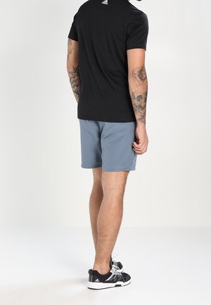 Mann trägt ein schwarzes Adidas-T-Shirt, graue Shorts und schwarze Adidas-Sneaker, steht mit dem Rücken zur Kamera, mit sichtbaren Tattoos an beiden Armen.