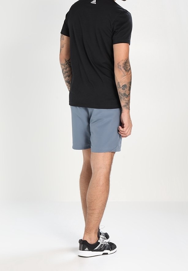 Homme portant un t-shirt Adidas noir, un short gris et des baskets Adidas noires, debout de dos, avec des tatouages visibles sur les deux bras.