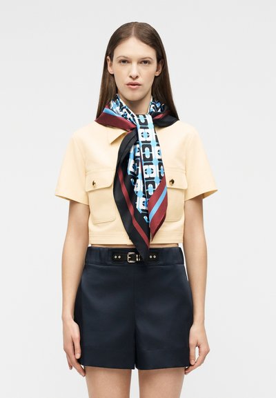 Γυναίκα με μπεζ πουκάμισο κοντομάνικο με τσέπες μπροστά, μεταξωτό κασκόλ με μπλε-κόκκινο-μαύρο μοτίβο και μαύρο σορτς ψηλόμεσο με ζωνάκι.