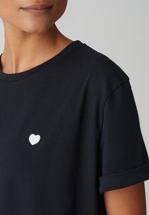Nahaufnahme einer Person, die ein schwarzes T-Shirt mit einem kleinen weißen Herz auf der linken Brust trägt.