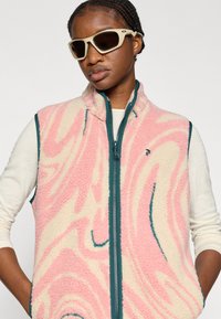 Γιλέκο από συνθετική γούνα με ροζ και κρεμ σχέδιο στροβιλίσματος, φερμουάρ και περιθώρια σε τιρκουάζ χρώμα. Φορεμένο πάνω από μακρυμάνικη κορυφή σε κρεμ απόχρωση. Αξεσουάρ περιλαμβάνουν αθλητικά γυαλιά ηλίου.