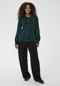 Donna sorridente con capelli ricci che indossa un blouse volanè verde scuro, pantaloni larghi neri e mocassini marroni, su uno sfondo semplice.