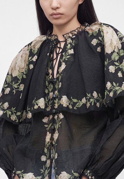 Жена, носеща прозрачна черна блуза с бежов флорален десен, надиплени ръкави, воланен якичка и закопчан с черен шнур отпред.