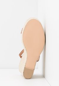 Sandale à semelle compensée beige avec côtés tissés, semelle lisse et bride à la cheville, positionnée semelle vers le haut contre un mur d’angle blanc.