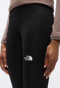 Μαύρο αθλητικό κολάν με ομαλή υφή και ελαστική μέση. Διαθέτει λευκό λογότυπο της "The North Face" στο μπροστινό μέρος του μηρού.