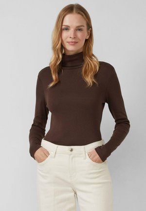 Blondynka z falowanymi włosami, ubrana w ciemnobrązowy golf i kremowe spodnie, stojąca z rękami w kieszeniach na jednolitym tle.